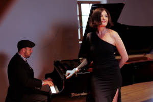 Eine Frau in schwarzem Kleid steht an einem Flügel, an dem ein Mann spielt