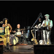 Eine Band aus einer Frau und vier Männern steht auf der Bühne. Die Frau singt, die Männer spielen Bass, Gitarre, Saxofon und Schlagzeug